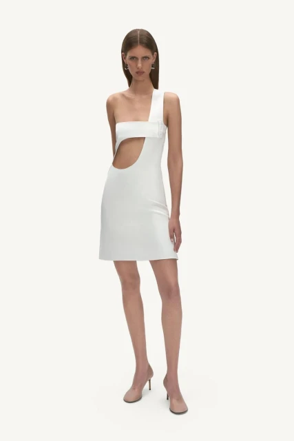 Full-length shot of a woman in a white one-shoulder mini dress with a cutout, standing in front of a white background. Nude-colored heels.