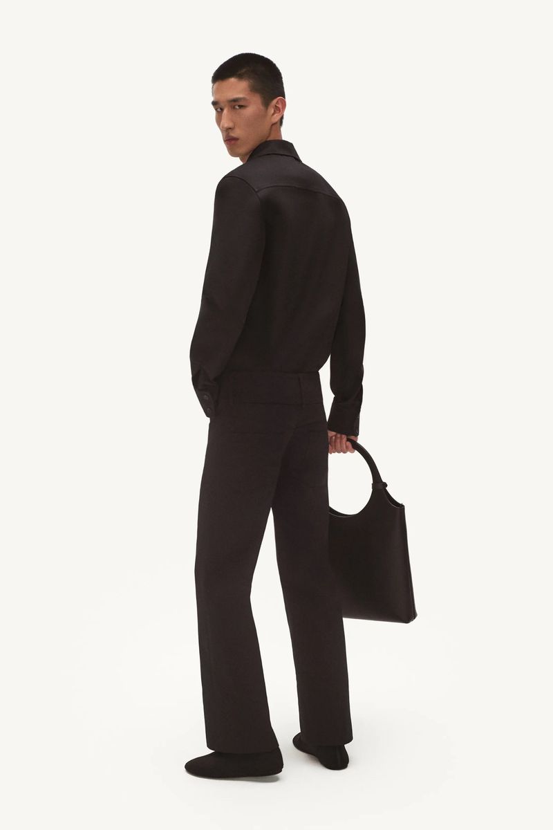 Man in black outfit with tote bag in studio. Man wearing black button-down shirt and flared black pants, holding large black tote.