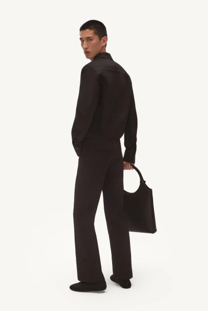 Man in black outfit with tote bag in studio. Man wearing black button-down shirt and flared black pants, holding large black tote.