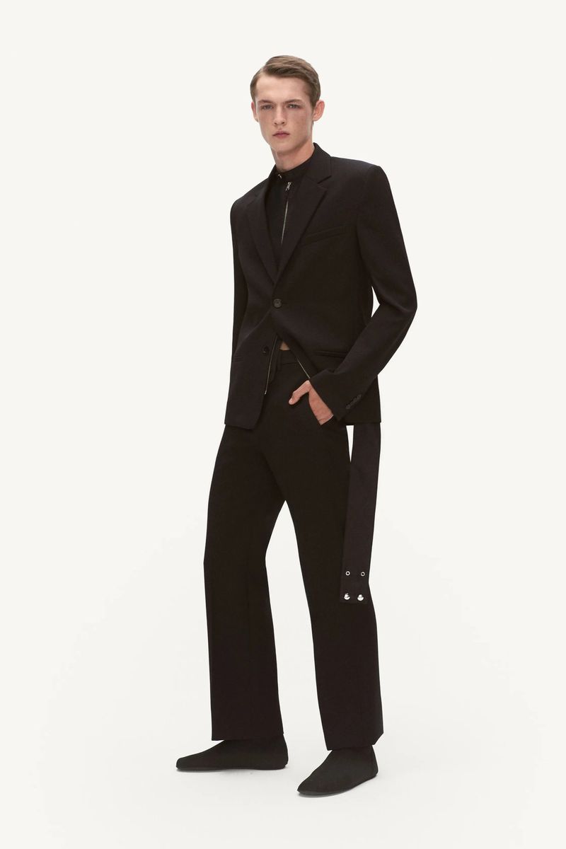 A male model poses in a black blazer, shirt, and pants outfit in a studio, exuding sleek fashion in a full-length, eye-level shot.