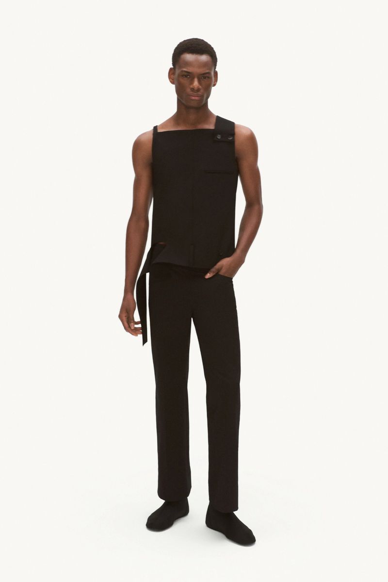Adult male stands in a full shot wearing a black top and pants. He's set against a white backdrop in a studio setting.