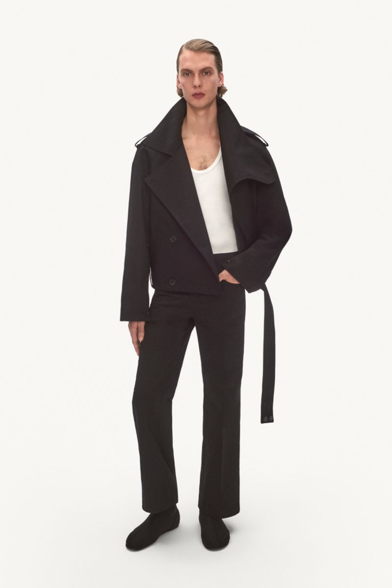 Full-length shot of adult male fashion model in black trench coat, white tank top, and black pants against a white backdrop in a studio.