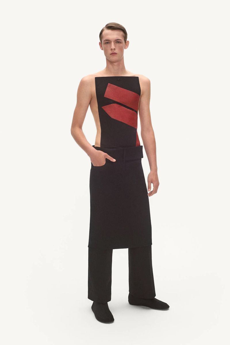 Young male fashion model in sleeveless top with red accents and draped pants in studio lighting, minimalist design, full-length shot.