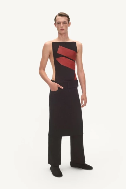 Young male fashion model in sleeveless top with red accents and draped pants in studio lighting, minimalist design, full-length shot.