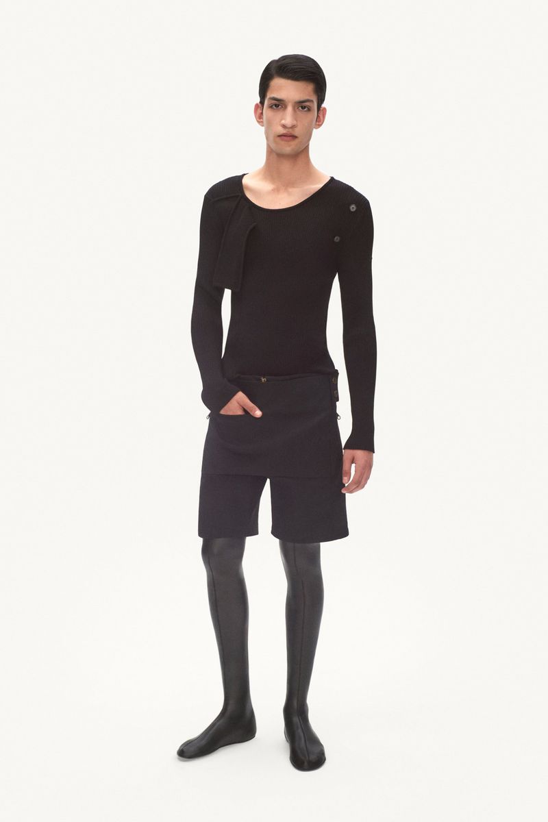 Model posing in an all-black fitted long-sleeve top, shorts with apron overlay, and tall knee-high boots on a white backdrop.