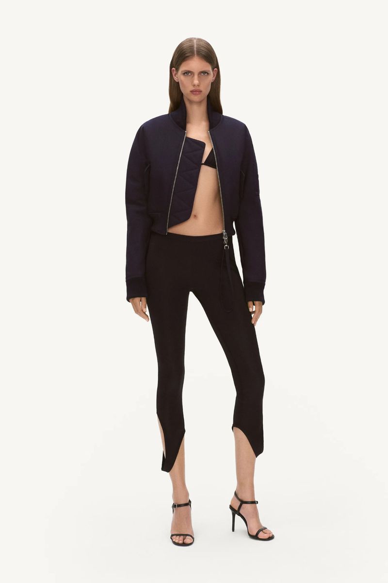 Fashion shot of a slim adult woman in a cropped navy bomber jacket, black bikini top, high-waisted leggings, and strappy high-heel sandals.