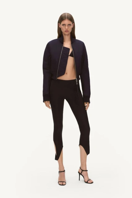 Fashion shot of a slim adult woman in a cropped navy bomber jacket, black bikini top, high-waisted leggings, and strappy high-heel sandals.