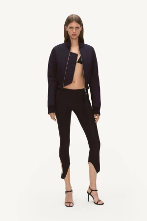 Fashion shot of a slim adult woman in a cropped navy bomber jacket, black bikini top, high-waisted leggings, and strappy high-heel sandals.