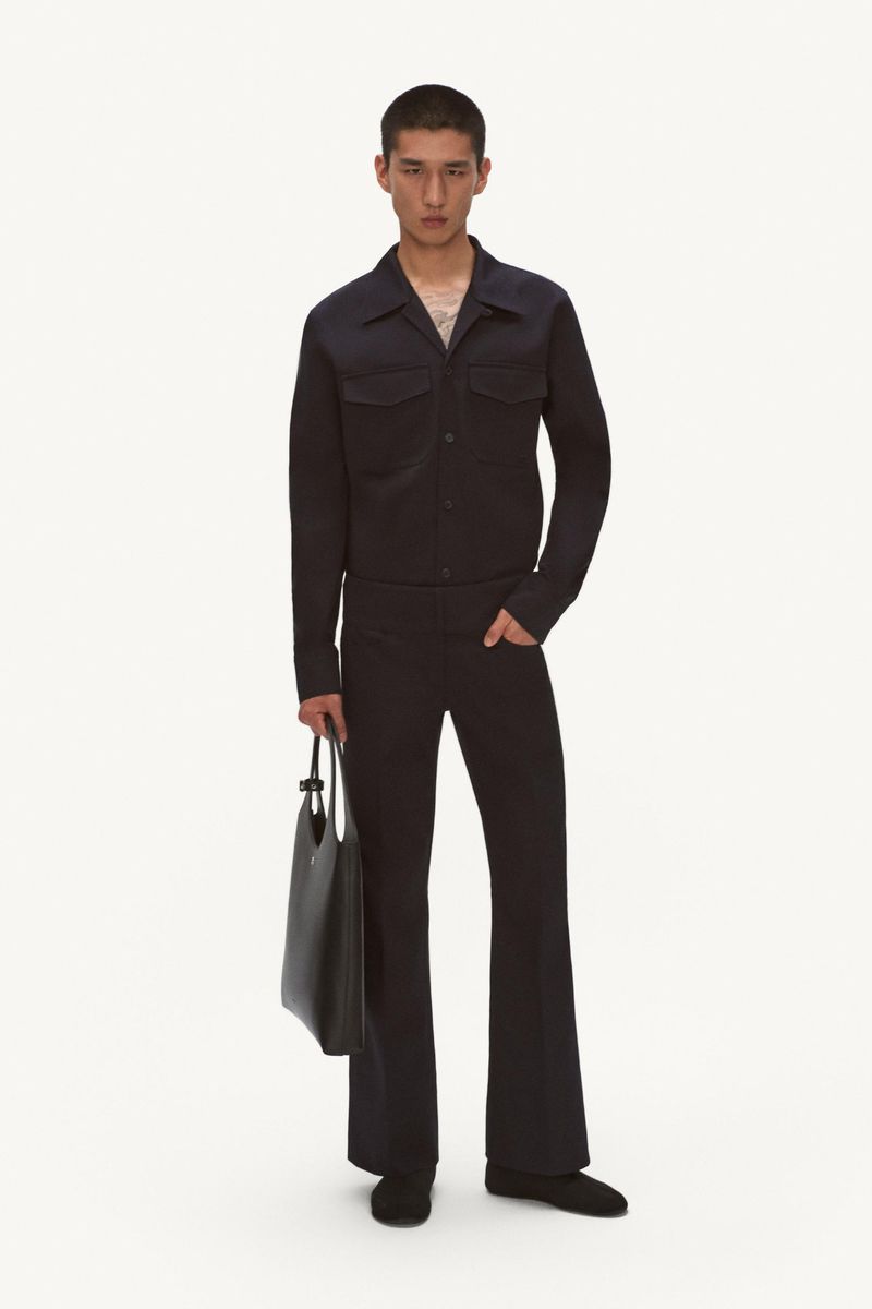 A young adult man poses in a dark blue collared jacket and flared trousers with a black tote bag on a white seamless background.