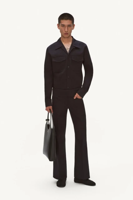 A young adult man poses in a dark blue collared jacket and flared trousers with a black tote bag on a white seamless background.