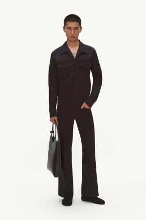 A young adult man poses in a dark blue collared jacket and flared trousers with a black tote bag on a white seamless background.