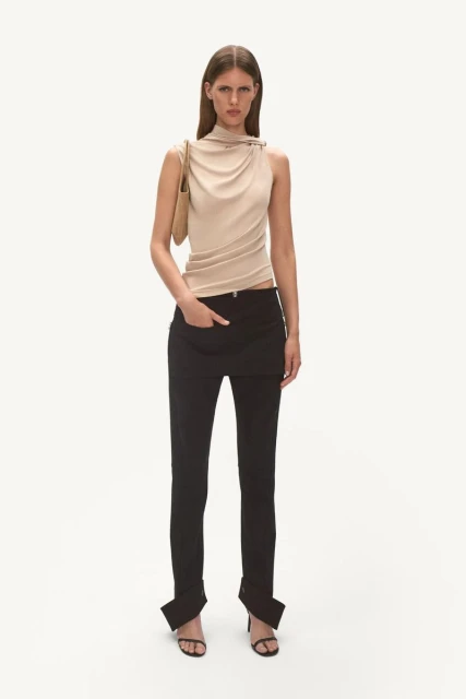 Full-length studio shot of a female fashion model in a draped top, black pants, heels, and a shoulder bag against a white backdrop.
