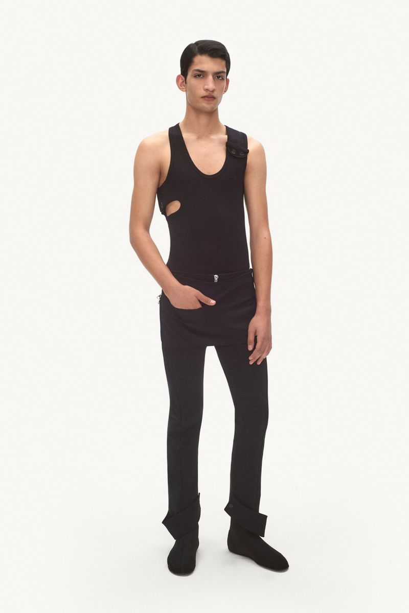 A slender man in a dark tank top and pants poses in a full shot, standing against a white background in a studio setting.