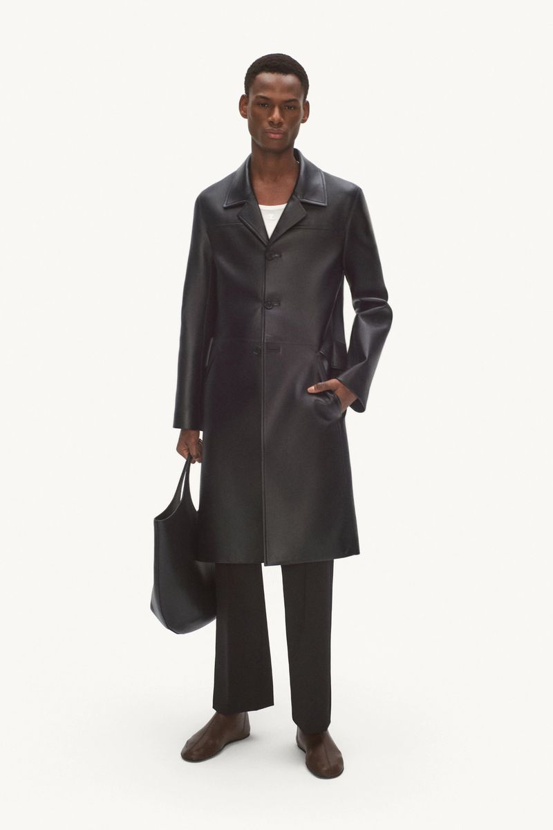 Full-length studio shot of an adult male wearing a black leather coat, black pants, and brown shoes, and holding a black tote bag.