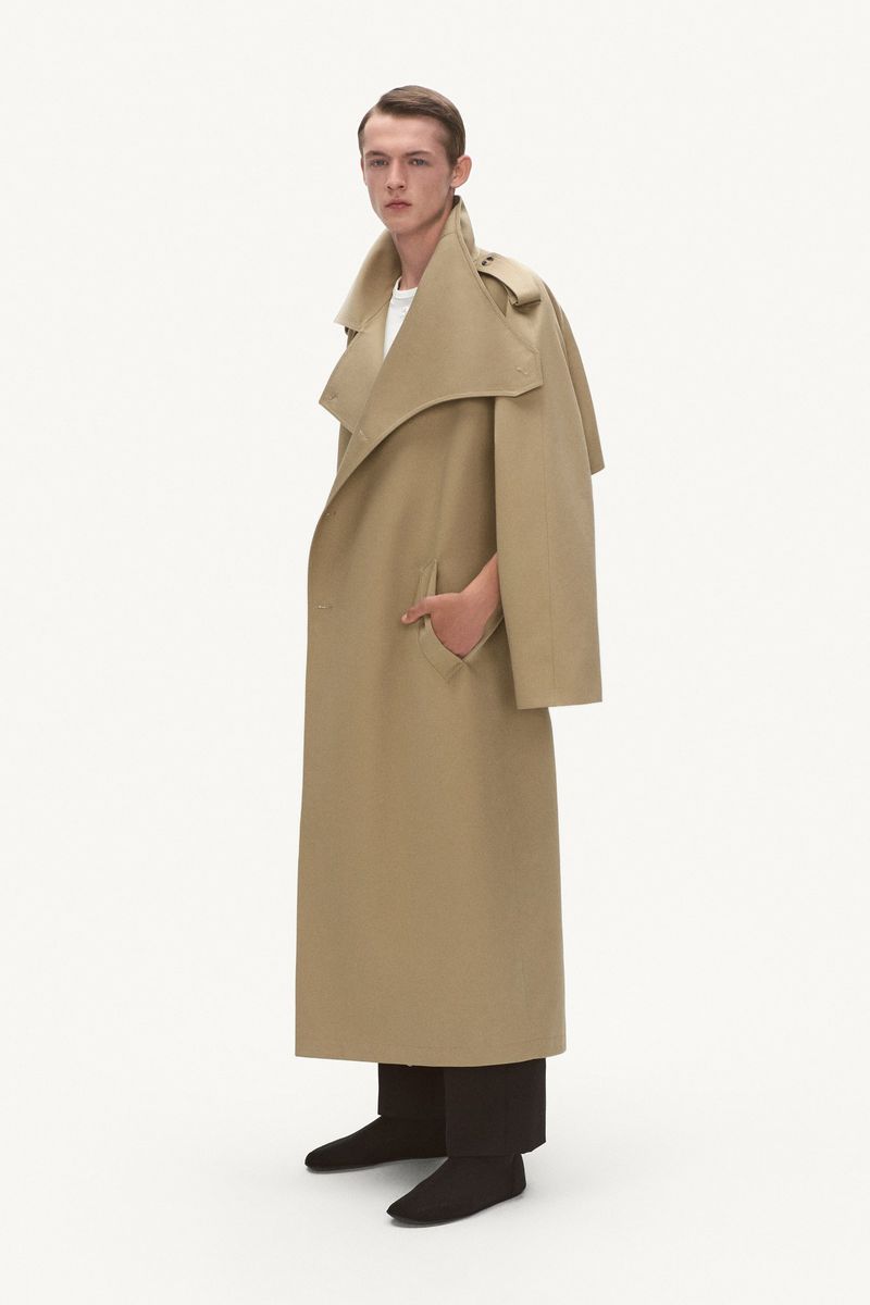 A slender man poses in a studio, wearing a long, beige trench coat with dark pants and shoes, against a plain white backdrop for a fashion shot.