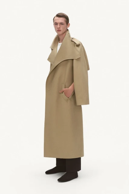 A slender man poses in a studio, wearing a long, beige trench coat with dark pants and shoes, against a plain white backdrop for a fashion shot.