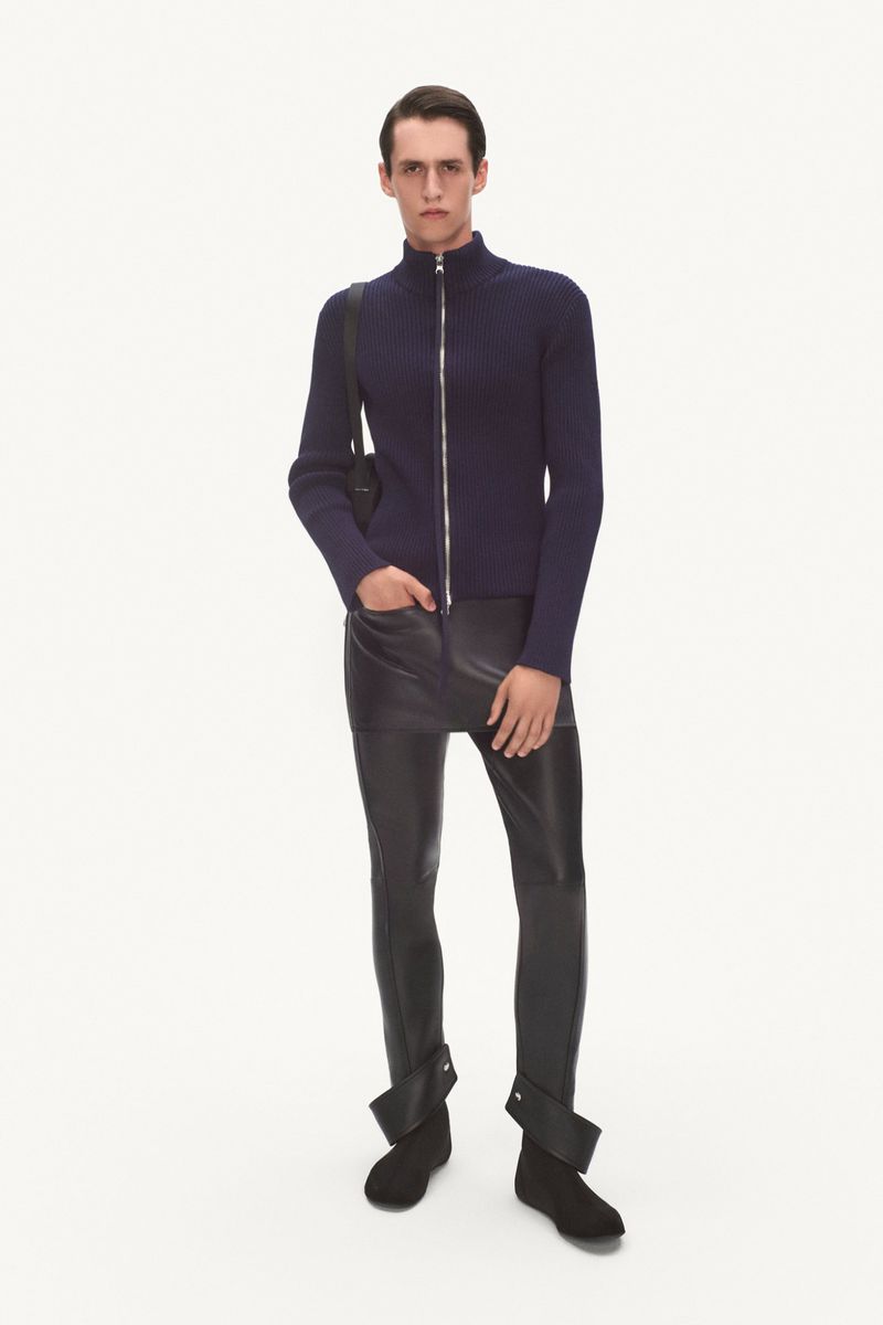 A stylish adult male in a navy ribbed zipper sweater, black leather-look leggings, and low boots against a bright white backdrop.