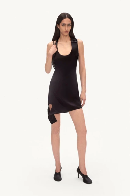 A slender fashion model poses in a fitted black mini dress and black high heels against a white backdrop in a studio setting.