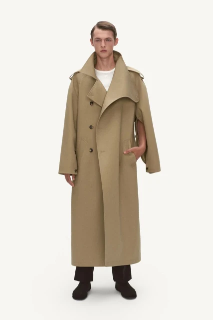 A full-length shot of a fair-skinned male model wearing a tan trench coat, white t-shirt, and black pants against a bright white backdrop.