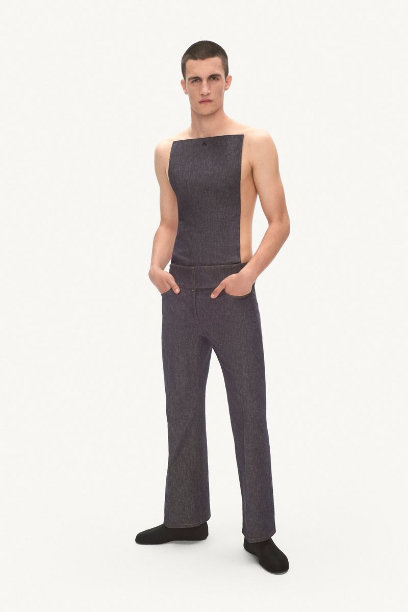 A young adult male posing in dark gray denim-like tunic and pants against a white backdrop in a studio setting, fashion shoot.