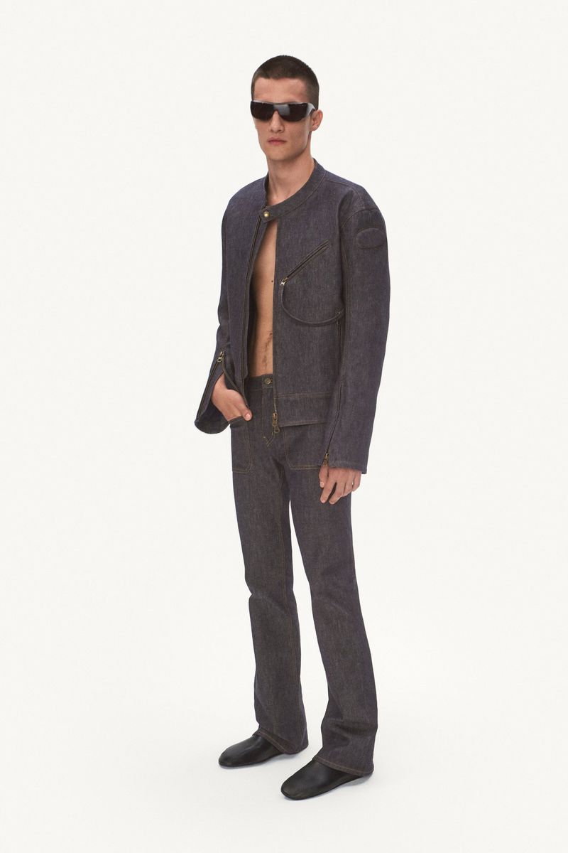 Model wearing a dark raw denim jacket, matching straight-leg trousers, and black sunglasses against a white studio background.