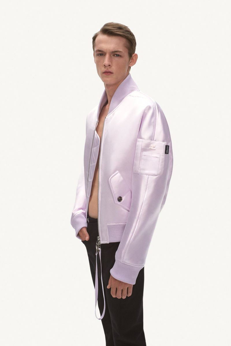 Young male model poses in a lilac bomber jacket and black pants against a stark white background in a fashion studio setting.
