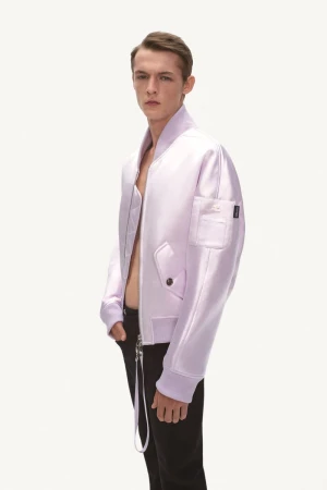 Young male model poses in a lilac bomber jacket and black pants against a stark white background in a fashion studio setting.