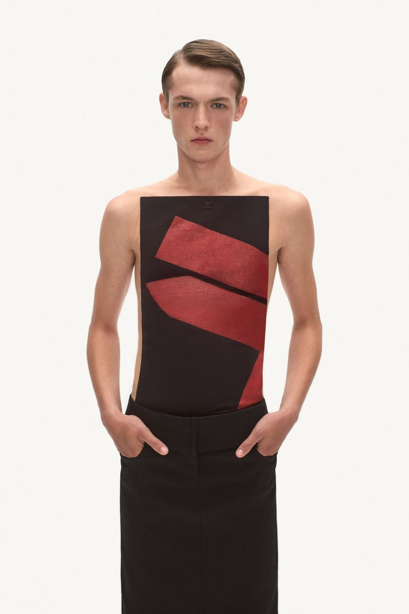 Fashionable adult male in a black top with red graphics and a long black skirt, hands in pockets, studio lighting and a neutral background.