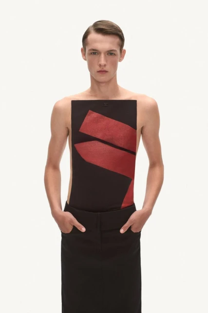 Fashionable adult male in a black top with red graphics and a long black skirt, hands in pockets, studio lighting and a neutral background.