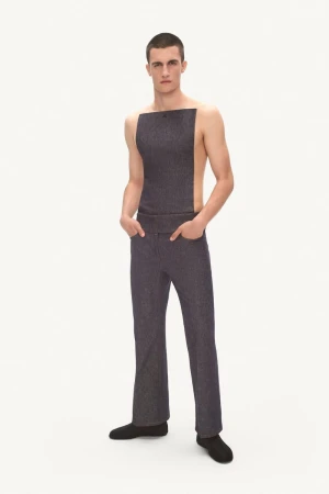 Model posing in a dark grey backless apron top and matching trousers with hands in pockets against a plain white backdrop.