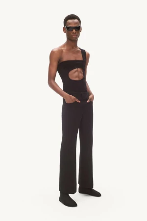 A Black male model stands in dark sunglasses, a black cutout one-shoulder top, and black trousers against a plain white studio background.