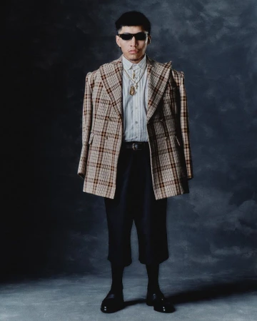 Man standing in an oversized plaid blazer, white shirt, dark sunglasses, layered gold necklaces, and cropped black trousers.