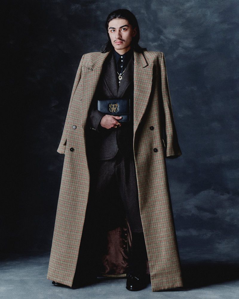 Portrait of a stylish adult male model with long hair, wearing a suit, long coat, and carrying a black clutch, against a dark backdrop.