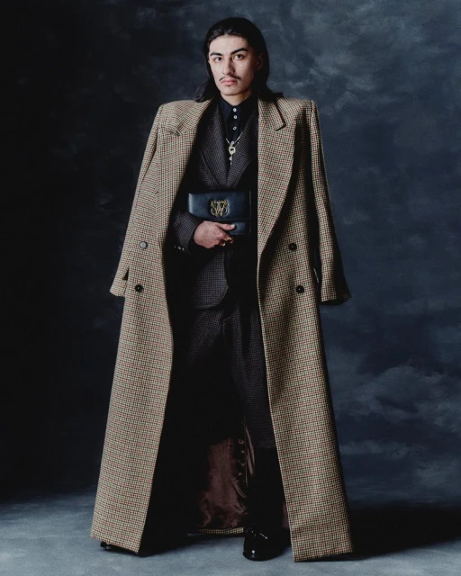 Portrait of a stylish adult male model with long hair, wearing a suit, long coat, and carrying a black clutch, against a dark backdrop.
