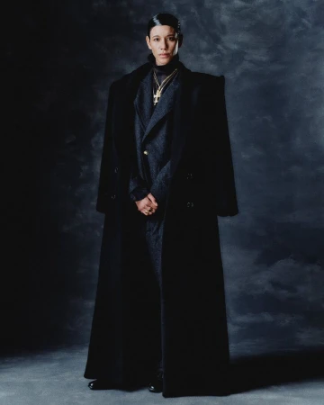 Model Dilone posing in a full-length black oversized coat and charcoal suit against a dark textured backdrop.