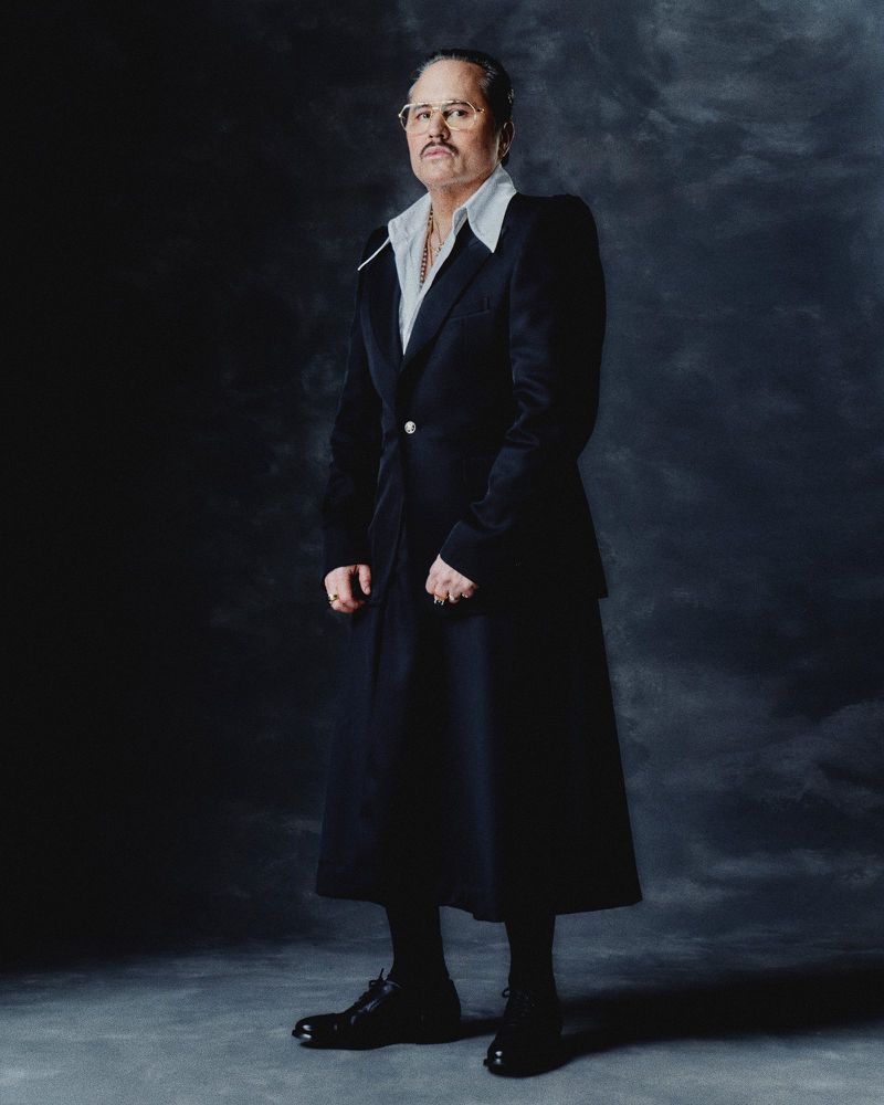 Studio portrait of a distinguished adult man in a tailored suit and glasses, standing against a textured dark gray background with soft lighting.