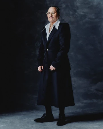 Studio portrait of a distinguished adult man in a tailored suit and glasses, standing against a textured dark gray background with soft lighting.