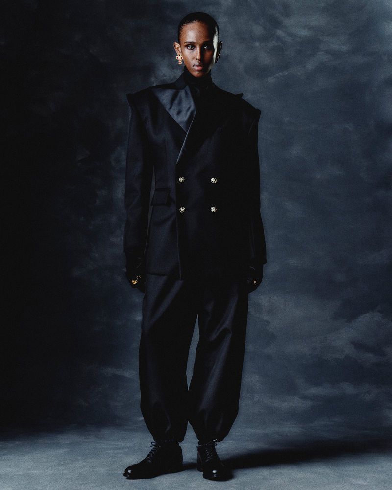 Fashion model in a stylish black suit with gold buttons and black boots stands in a dark photo studio, posing for an editorial shot.