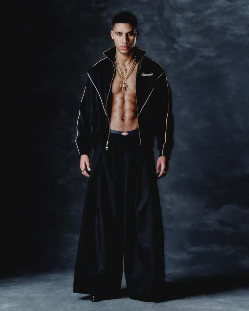 Man in black tracksuit and wide-leg pants poses against dark backdrop, showcasing fashion style and confident expression, captured in studio lighting.