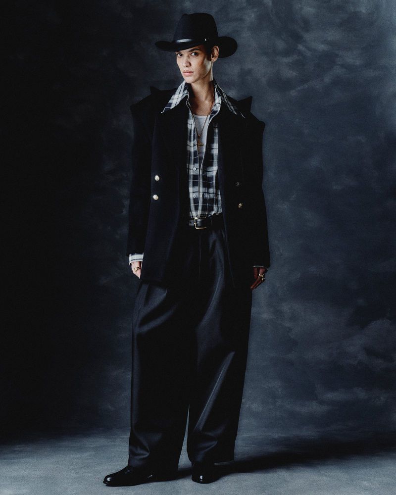A person stands wearing a cowboy hat, blazer, and wide-legged pants against a dark backdrop with shadows, looking forward in a fashion pose.