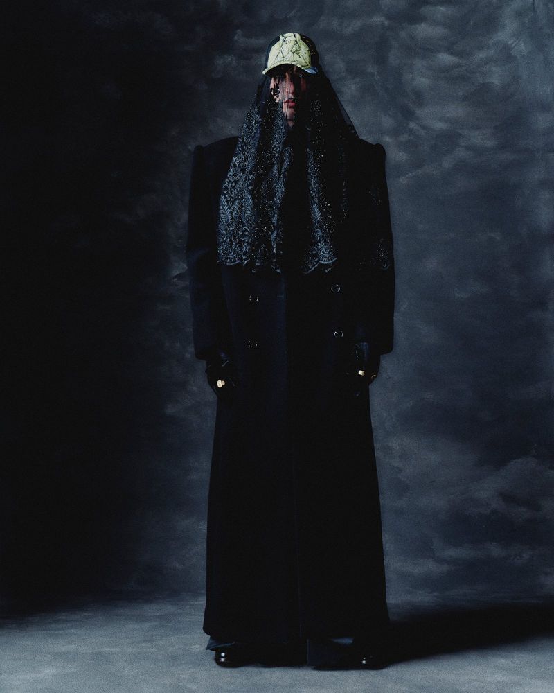 Full body shot of a person wearing a baseball cap and lace veil over their head, a long coat, and black shoes against a dark backdrop.
