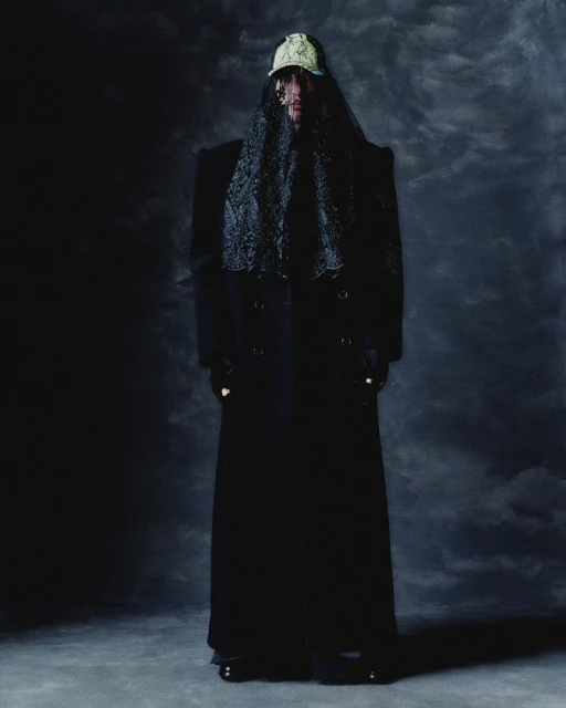 Full body shot of a person wearing a baseball cap and lace veil over their head, a long coat, and black shoes against a dark backdrop.