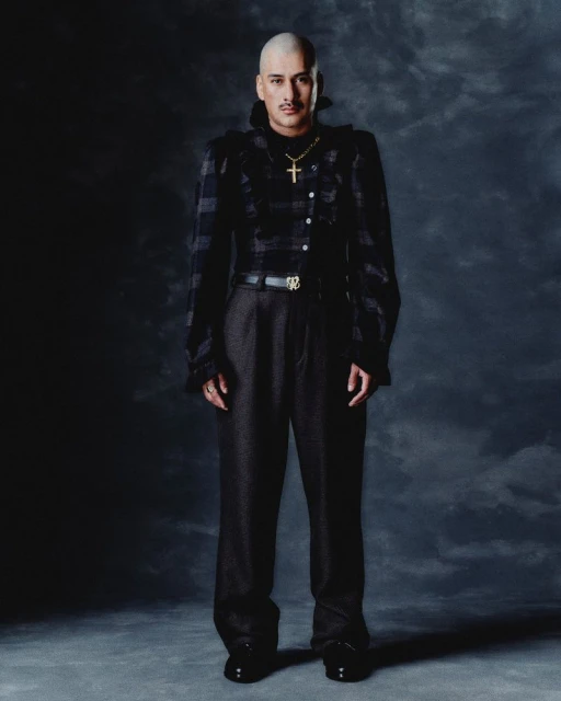 Bald adult male in a black plaid jacket, gold cross necklace, dark trousers and shiny shoes stands against a mottled dark backdrop.