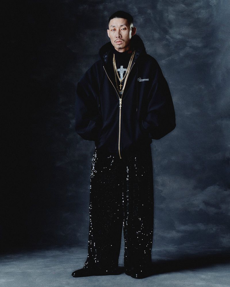 Portrait of an adult male in a black hooded jacket, black pants, and jewelry, posing in a studio with a dark backdrop.