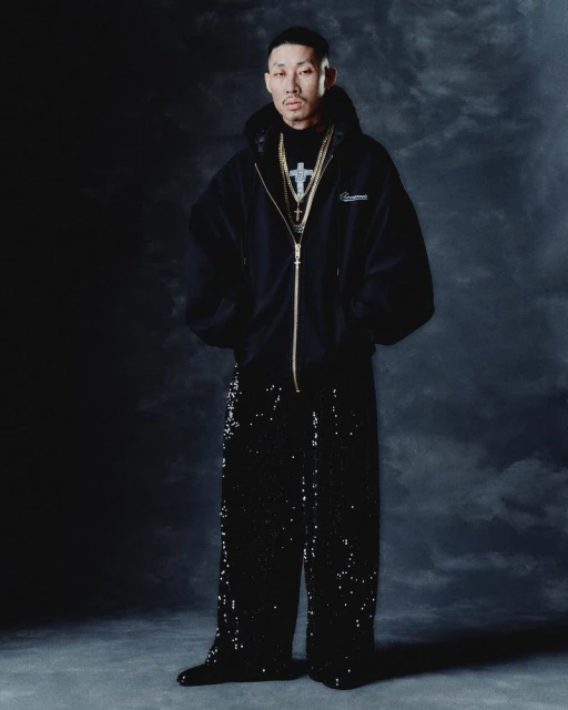 Portrait of an adult male in a black hooded jacket, black pants, and jewelry, posing in a studio with a dark backdrop.