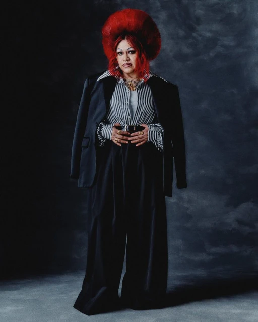 Full-length portrait of a person with red hair, wearing black pants, a striped shirt, and a black blazer, set against a dark background.