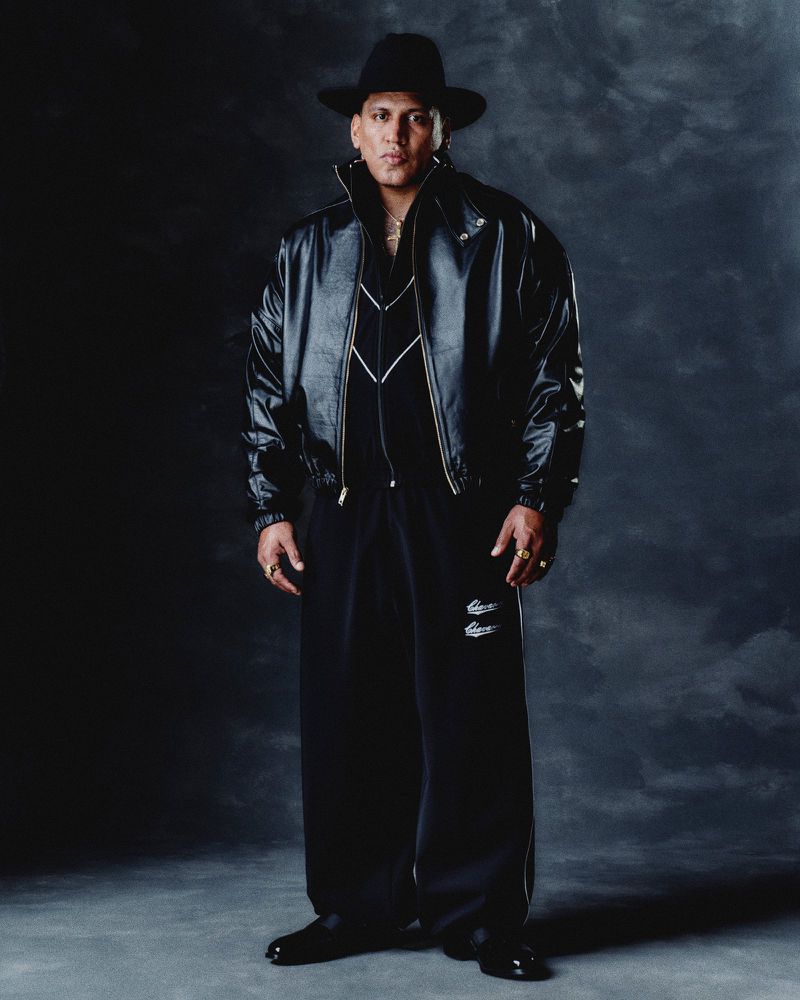 Portrait of a single adult male in a black leather jacket, hat, and sweatpants standing in a photo studio with dark background.
