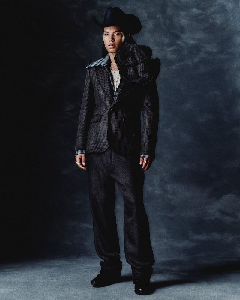 Male fashion model wearing a dark suit and cowboy hat poses in a studio with a textured background, showcasing modern Western style.