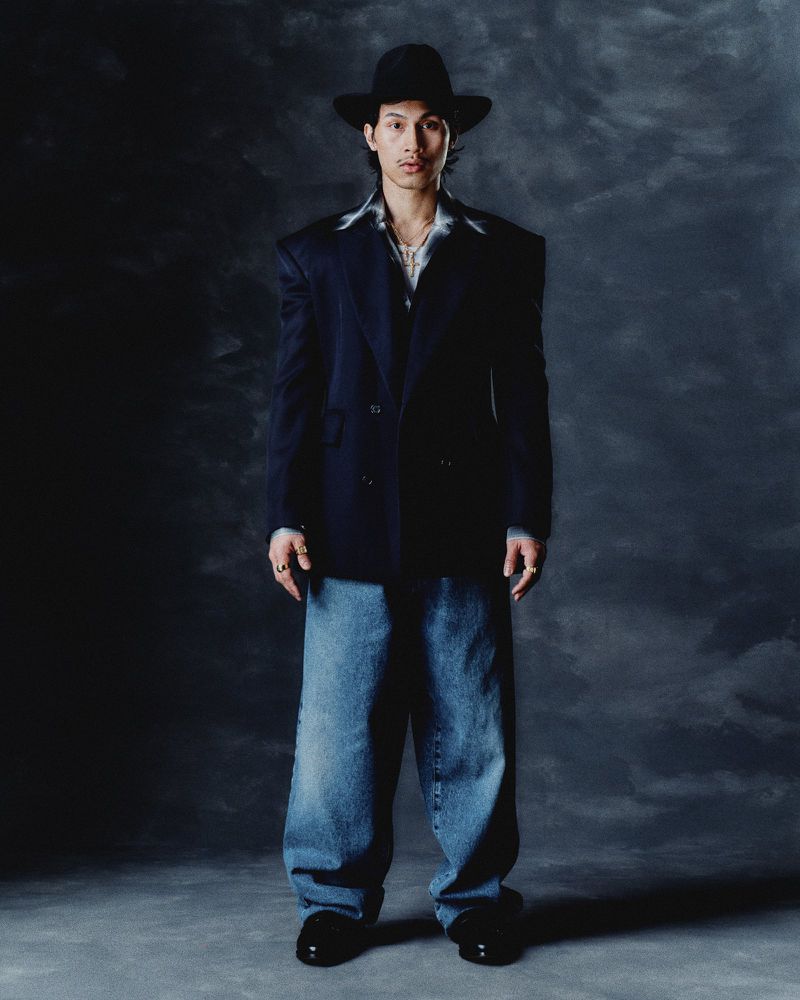 Portrait of a young man in a black hat, oversized blazer, and baggy jeans against a mottled dark gray backdrop in a fashion shot.