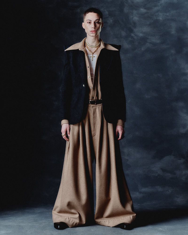 Full-length studio shot of a person wearing a black jacket, beige shirt, tan pants, jewelry, and black shoes against a dark backdrop.